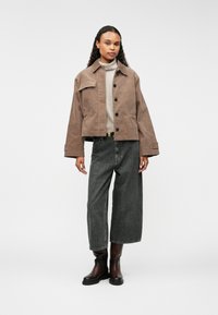 Chaqueta de pana marrón con cuello, bolsillo frontal y cierre de botón, llevada sobre un suéter gris y combinada con jeans de pierna ancha oscuros y botas marrones.