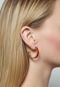 Goldene Creole mit einem glatten roten Akzent, die ein modernes, offenes Design aufweist. Die Haare sind glatt frisiert und verdecken teilweise das Ohr.