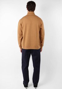 Pull en polaire marron avec col montant, doté d'un ourlet côtelé. Porté avec un pantalon bleu marine et des chaussures noires. Texture lisse, design décontracté.