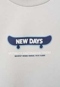 Felpa grigia chiara con grafica che presenta testo blu e bianco: "NUOVI GIORNI" e "TRUCCHI DI SKATEBOARD FINALE" con illustrazione di skateboard.