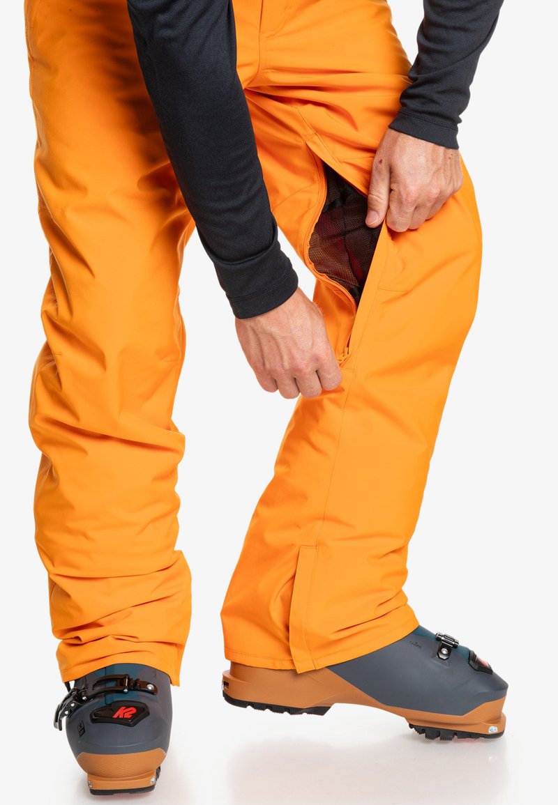 Pomarańczowe spodnie narciarskie z zapięciem po bokach, noszone z szarymi butami narciarskimi z czarnymi elementami. Materiał wydaje się gładki i trwały.