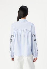Camisa azul-clara de botão com mangas longas, apresentando bordado floral preto nas mangas e detalhe franzido nas costas. Tecido de algodão suave.
