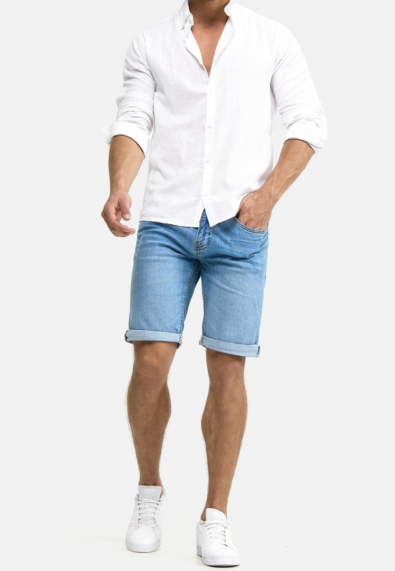 Chemise blanche en lin avec boutons, short en denim bleu clair à ourlet retroussé, baskets blanches. Tenue décontractée avec une coupe ample et des couleurs claires.