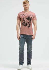 Pink cotton t-shirt with a black graphic print, v-neck, and short sleeves, paired with light blue denim jeans and white sneakers.