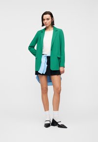 Femme portant un blazer vert, un haut blanc, une jupe noire, une chemise bleu clair nouée à la taille, des chaussettes blanches et des chaussures à talons noires, debout devant un fond uni.
