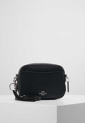 Bolso cruzado Coach de cuero negro con logo plateado, bolsillo frontal, cremallera con borla y correa desmontable sobre pedestal blanco.