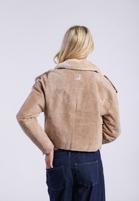Veste en daim beige avec un col en fourrure, design cropped et épaulettes ; la vue arrière révèle une texture douce et un patch logo. Jean en denim foncé.