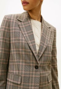 Blazer à carreaux dans des tons gris, beige et rouge. Il présente un col cranté, une fermeture à un bouton et deux poches plaquées à l'avant. Tissu texturé.