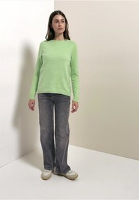 Pull en tricot vert clair à col rond, associé à un jean en denim gris et des baskets beiges, debout devant un fond neutre.