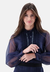 Navy blouse with sheer sleeves, ruffled front, and a decorative trim. Features buttoned cuffs and a textured waistline.