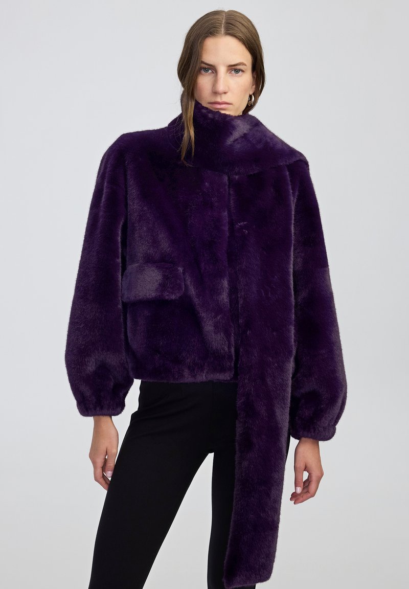 Donna con una giacca in pelliccia sintetica di un viola scuro con una lunga sciarpa attaccata, abbinata a pantaloni neri, su uno sfondo neutro.