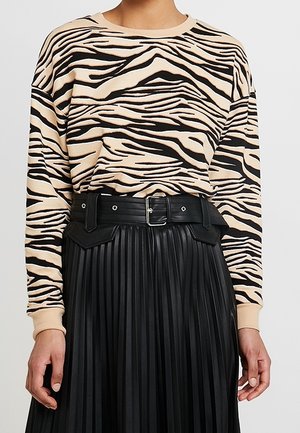 Kobieta ubrana w beżowo-czarną bluzkę w paski zebry z długim rękawem, wpuszczoną w wysokowatą czarną plisowaną spódnicę z szerokim paskiem.
