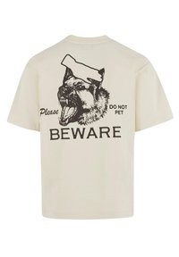 Cremefarvet bomulds T-shirt med grafik af en gøende hund iført en hat, med teksten "Venligst VÆR OBS på - MÅ IKKE KÆLES" i sort.