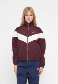 Burgundi ja valge värvi blokeeritud jakk kõrgema kraega, täis tõmblukuga ja elastsete mansettidega. Rinnal on Nike logo. Pehme, tekstuuriga kangas.