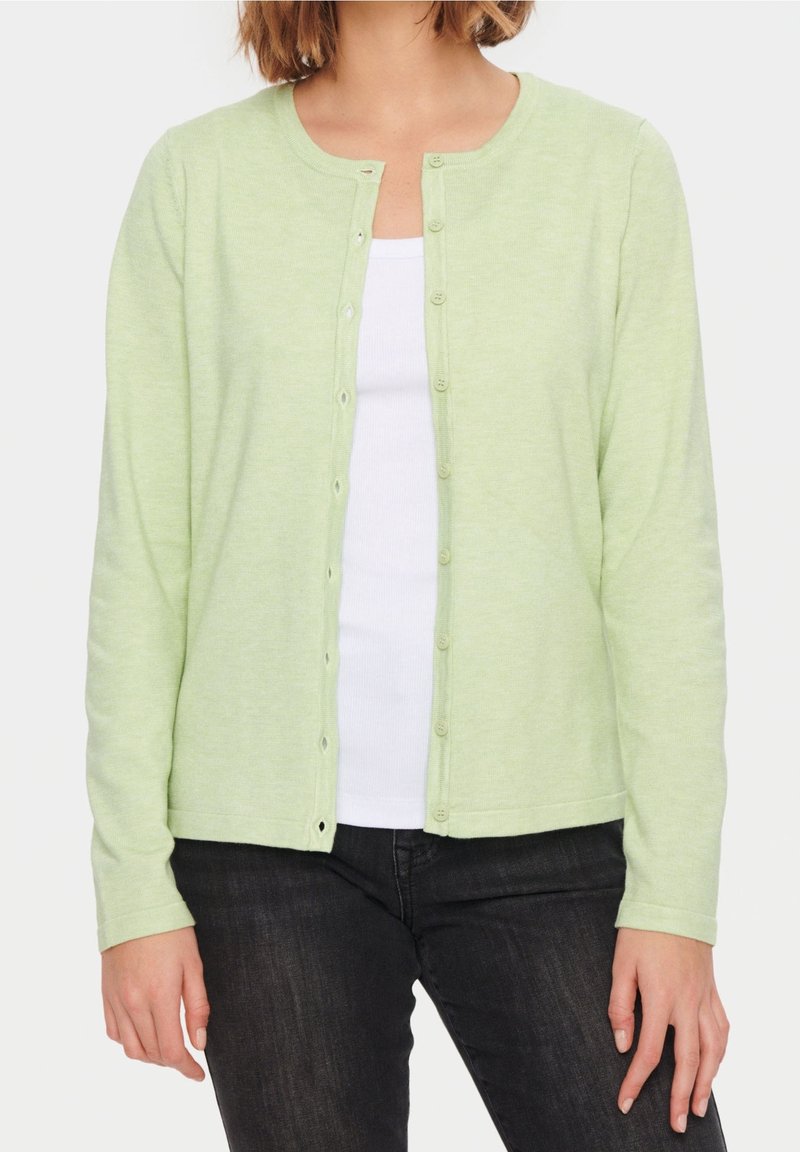 Femme portant un cardigan vert clair boutonné par-dessus un haut blanc et un jean foncé, debout les mains détendues le long du corps.