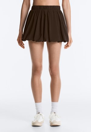 Nainen pukeutuneena lyhyeen, tummanruskeaan kuplahameeseen, jossa elastinen vyötärönauha, valkoiset sukat ja beige-valkoiset tennarit, seisoo yksiväristä taustaa vasten.