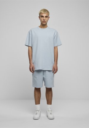 Junger Mann mit kurzem blondem Haar steht frontal und trägt ein hellblaues, übergroßes T-Shirt sowie passende Shorts mit weißen Socken und Turnschuhen.