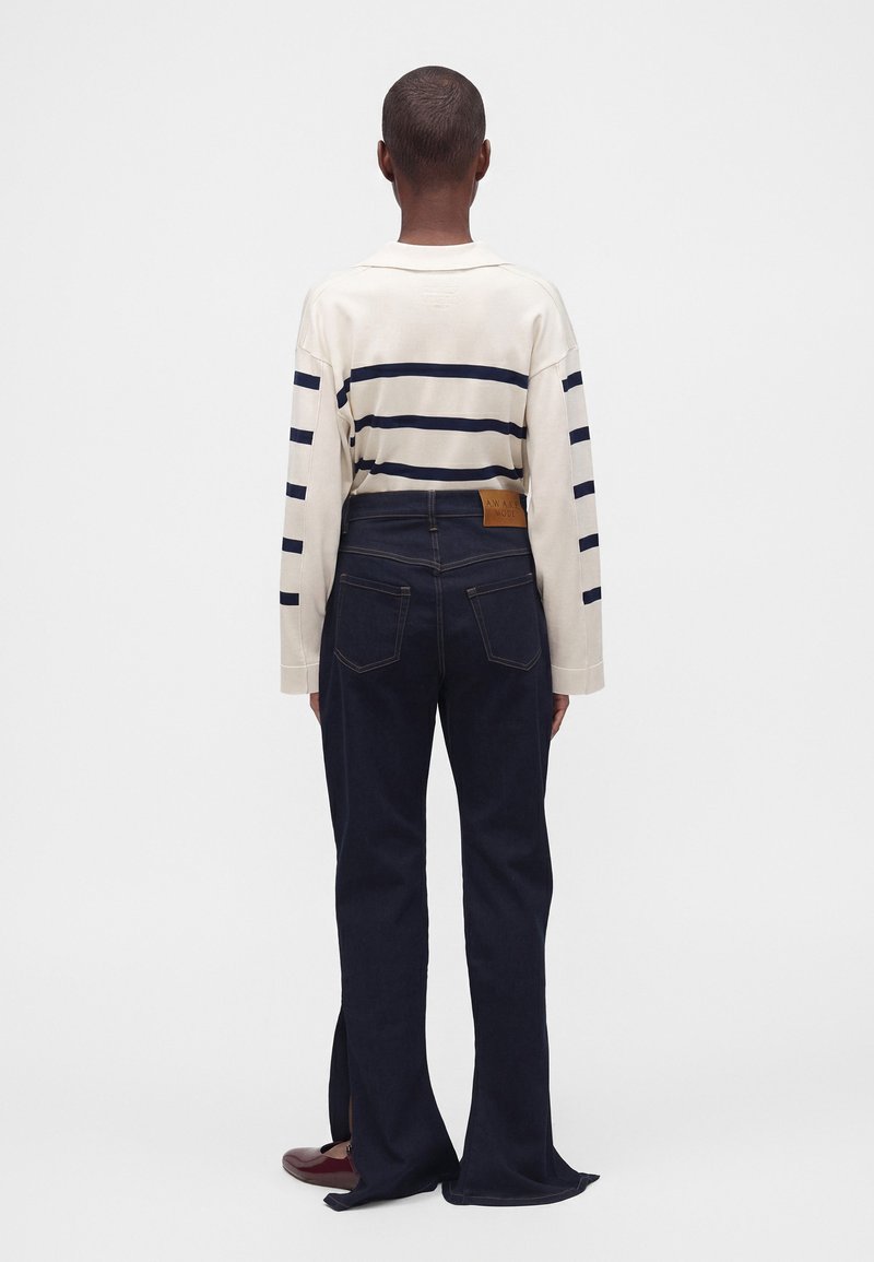 Personne aux cheveux courts, vue de dos, portant une chemise crème à manches longues avec des rayures bleu marine et un jean large bleu foncé avec des ourlets fendus.