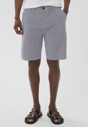 Mężczyzna noszący czarne skórzane sandały i szare szorty w pionowe białe paski, z rękami w kieszeniach na białym tle.