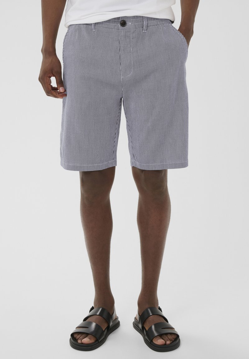 Homme portant des sandales en cuir noir et un short à rayures verticales grises et blanches, les mains dans les poches, sur un fond blanc.