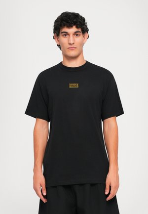 Young man with curly hair wearing a loose black t-shirt with a small rectangular gold logo patch on the chest, standing against a plain white background.