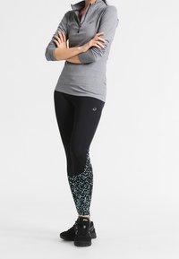 Grauer Reißverschluss-Sportoberteil, schwarze Leggings mit blauem gepunktetem Muster an der Unterschenkelpartie, kombiniert mit schwarzen Sneakern.