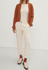 rostorange Strickjacke, cremiges Oberteil mit linearer Textur, hellbeige Cordhosen und blasse Slipper über braunen Socken.