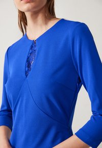 Robe bleue avec des manches trois-quarts, présentant un détail en dentelle au niveau du col, et un design ajusté avec une texture subtile.