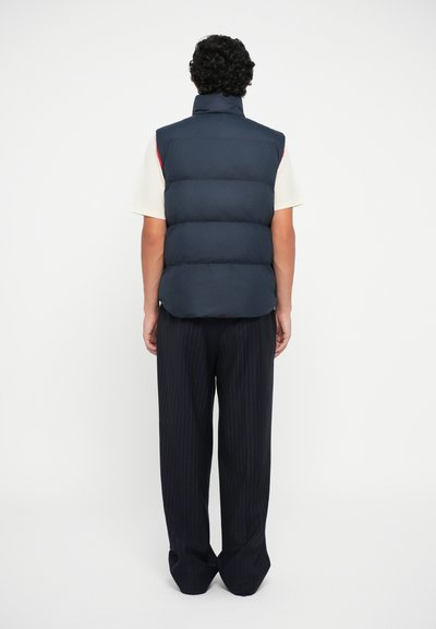 Navy puffer vest with high collar, featuring horizontal quilted pattern. Worn over a light shirt and paired with pinstriped trousers.
