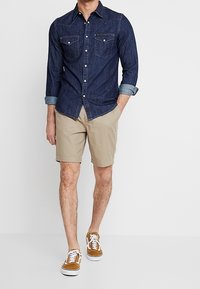 Czarna dżinsowa koszula z długim rękawem i dwoma kieszeniami na piersi, khaki szorty i brązowe trampki z białą podeszwą. Swobodny strój z podwiniętymi rękawami.