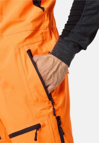 Felle oranje werkoveralls zijn voorzien van meerdere ritszakken, contrasterende zwarte accenten en gestructureerde zwarte mouwen voor extra duurzaamheid en functionaliteit.
