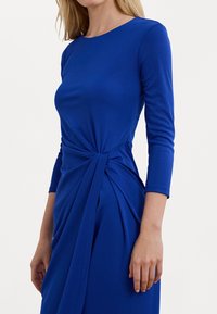 Blå långärmad klänning i ett mjukt material, med en snodd detalj i midjan och samlad tyg på sidan.