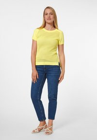 Geel mesh T-shirt met korte mouwen, gecombineerd met donkerblauwe skinny jeans en witte sandalen met bandjes. Het T-shirt heeft een ronde halslijn en zijsplits.