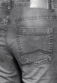 Jean en denim gris avec une texture décolorée, présentant une poche arrière unique avec une couture courbée et une étiquette près de la taille, détails de couture visibles.