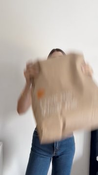 Bruine papierenzak met een gladde textuur, met een klein oranje logo en witte tekst, vastgehouden door een persoon in casual kleding tegen een lichte achtergrond.