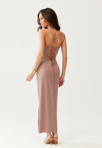 Žena v dlouhé fialové maxi šatech bez rukávů s křížovým šněrováním na zádech, se zlatým náramkem a béžovými vysokými podpatky, stojící na bílém pozadí.