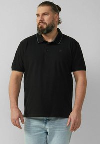 Schwarzes Polohemd mit kurzen Ärmeln, versehen mit einem kontrastierenden Kragen und Knöpfen. Kombiniert mit hellblauen Jeans. Dezentes Logo auf der Brust.