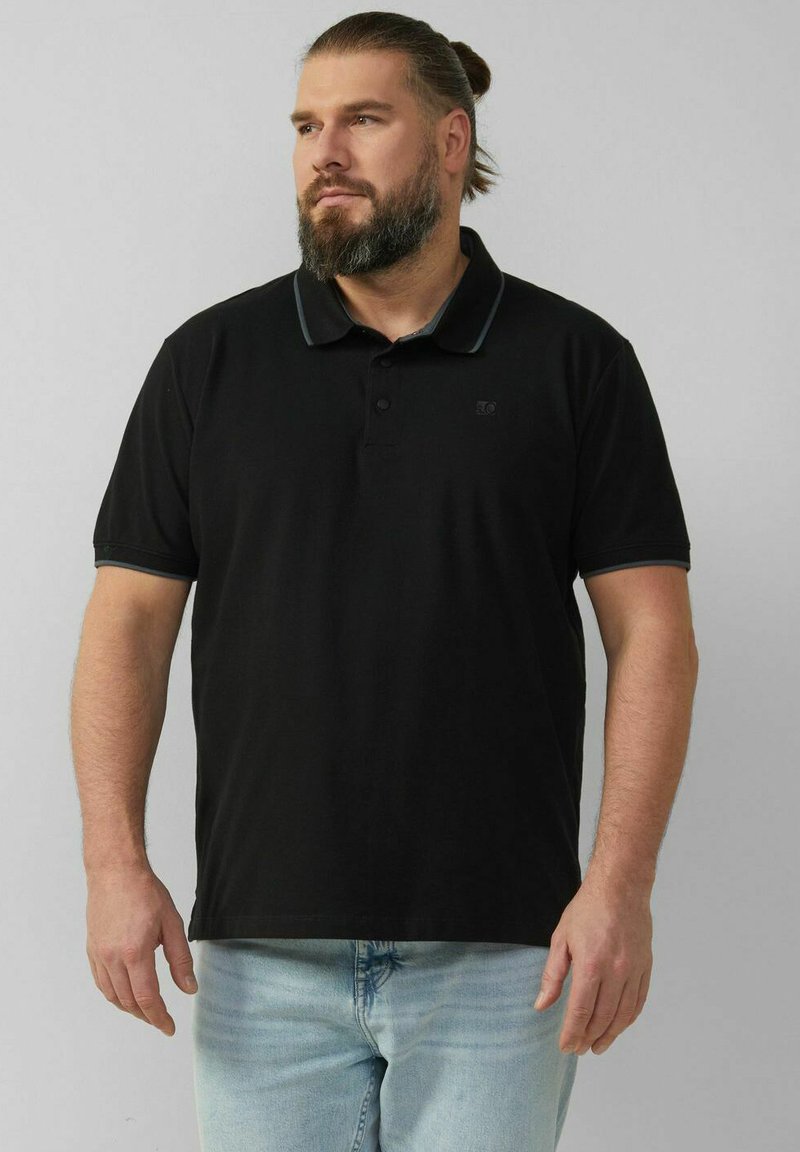 Schwarzes Polohemd mit kurzen Ärmeln, versehen mit einem kontrastierenden Kragen und Knöpfen. Kombiniert mit hellblauen Jeans. Dezentes Logo auf der Brust.