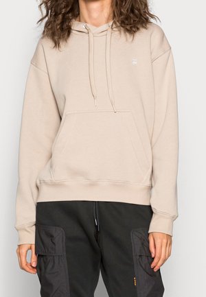 Osoba ubrana w beżową bluzę z kapturem i kieszenią z przodu oraz czarne spodnie z dużymi bocznymi kieszeniami.