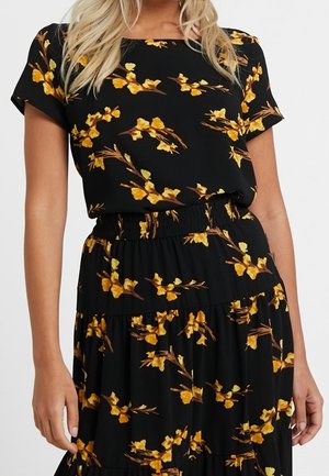Vrouw die een zwarte jurk met korte mouwen en geel bloemmotief draagt, een elastische tailleband en een geplooide rok.