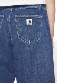 Blå denimjeans med en firkantet baklomme som har et hvitt Carhartt-logo merkelapp. Stoffet har en teksturert overflate med synlige sømmer.