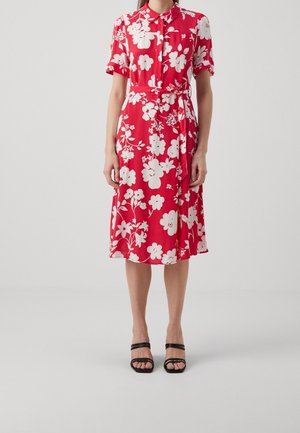 Rode bloemenjurk met korte mouwen, knopen aan de voorkant en een tailleband. Heeft witte bloemenprints en een A-lijn rok die tot de knie reikt. Zwarte sandalen met bandjes.