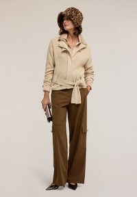 Beige žebrovaný svetr s páskem, hnědé široké kalhoty a klobouk s leopardím potiskem. Metalická kabelka a černé špičaté boty.