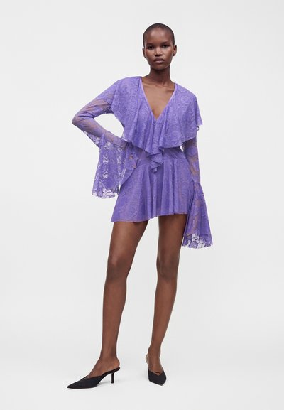 Femme debout, main sur la hanche, portant une robe en dentelle violette avec un décolleté en V profond, des manches évasées, une jupe plissée et des talons noirs à bout pointu.