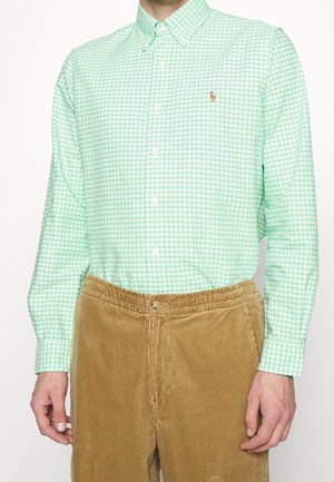 Homme portant une chemise à carreaux vert menthe et blanc à boutons et un pantalon en velours côtelé marron clair, debout sur fond blanc.