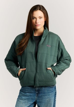 Vrouw draagt een groene doorgestikte jas met voorzakken en blauwe spijkerbroek, staand tegen een eenvoudige lichte achtergrond.