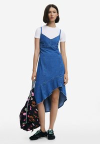 Denim dress with a high-low hem, spaghetti straps, and contrast stitching. Paired with a floral bag and black sneakers with turquoise accents.