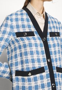 Cardigan a quadri blu e bianchi con scollo a V, bordature nere e due tasche frontali, caratterizzato da bottoni lucidi e un tessuto testurizzato.