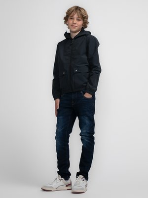 Černá kapucínová bunda se zipem vpředu, elastickými manžetami a dvěma předními kapsami; v kombinaci s tmavými džíny a bílými teniskami s červenými akcenty.