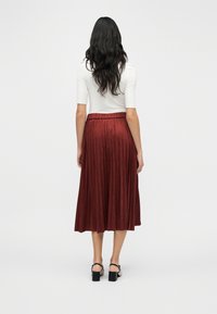 Μίν τι φούστα με πιέτες σε βαθύ κόκκινο, κατασκευασμένη από μαλακό ύφασμα. Διαθέτει μέση με ζώνη και λεία υφή. Φοριέται με λευκό τοπ και μαύρα λουράκια στον αστράγαλο.